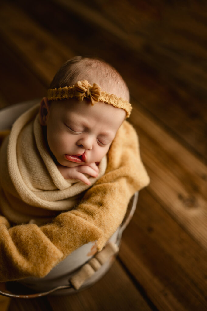 Baby girl wrapped in yellow with a yellow head band. She is posed in a buck with her head resting against the side of the bucket during her newborn session