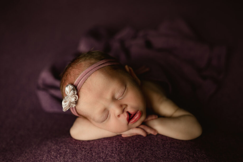 Baby girl posed with hands on chin on a purple backdrop during her newborn session