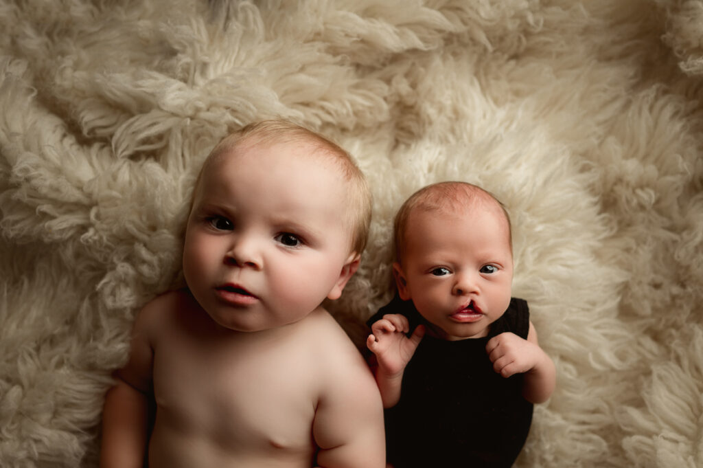 Big brother and baby sister posed laying on their backs side by side on a fuzzy nude rug.