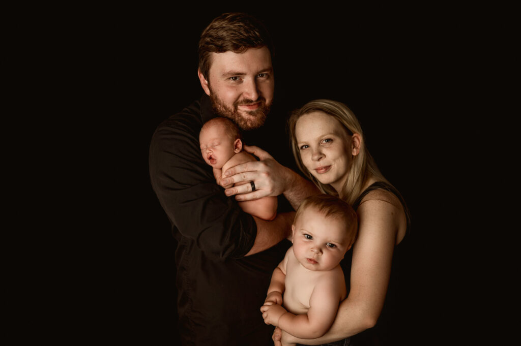 Dad holding baby and mom holding older brother standing side by side on a solid black backdrop