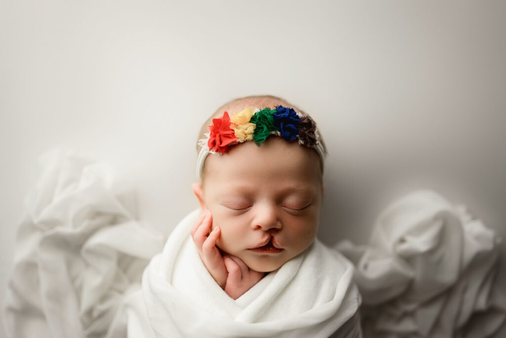 Baby girl wrapped in white with a white back drop and white layering wrap wrapped over her mid section. Baby is wearing a headband with rainbow colored bows during her newborn session