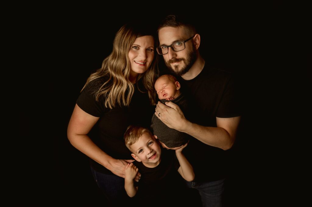 Family of four posed for a newborn portrait session on a black backdrop. 