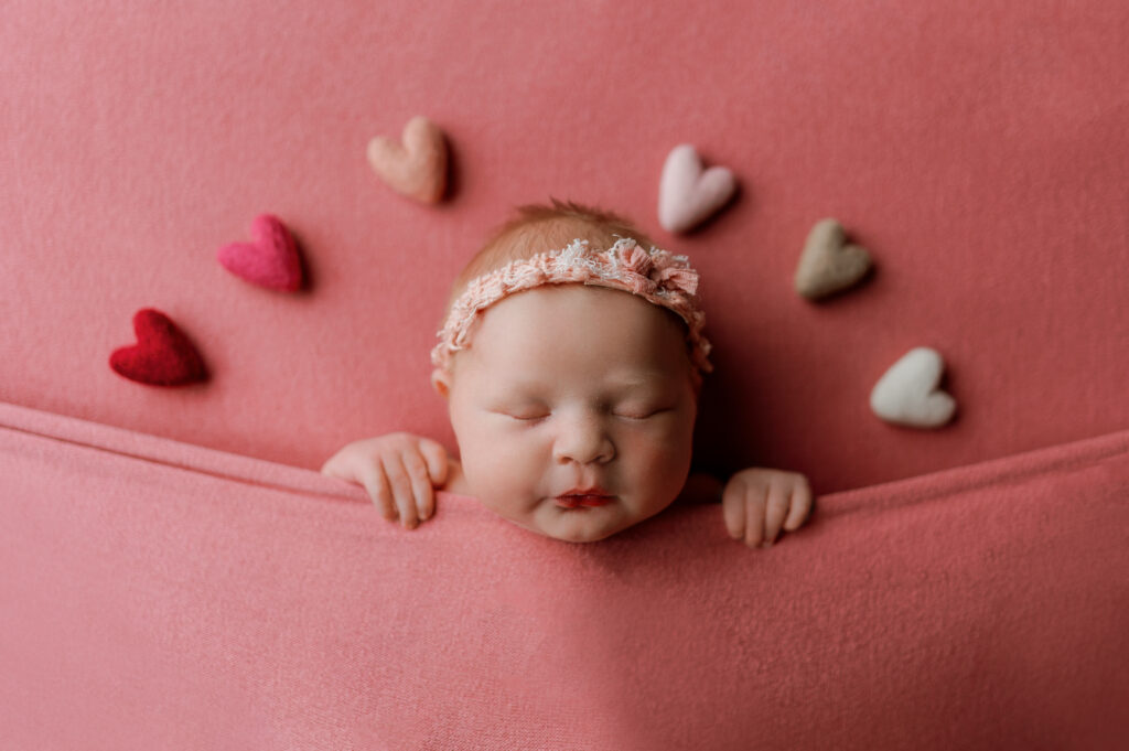Baby on pink backdrop tucked in with hearts above her head. 