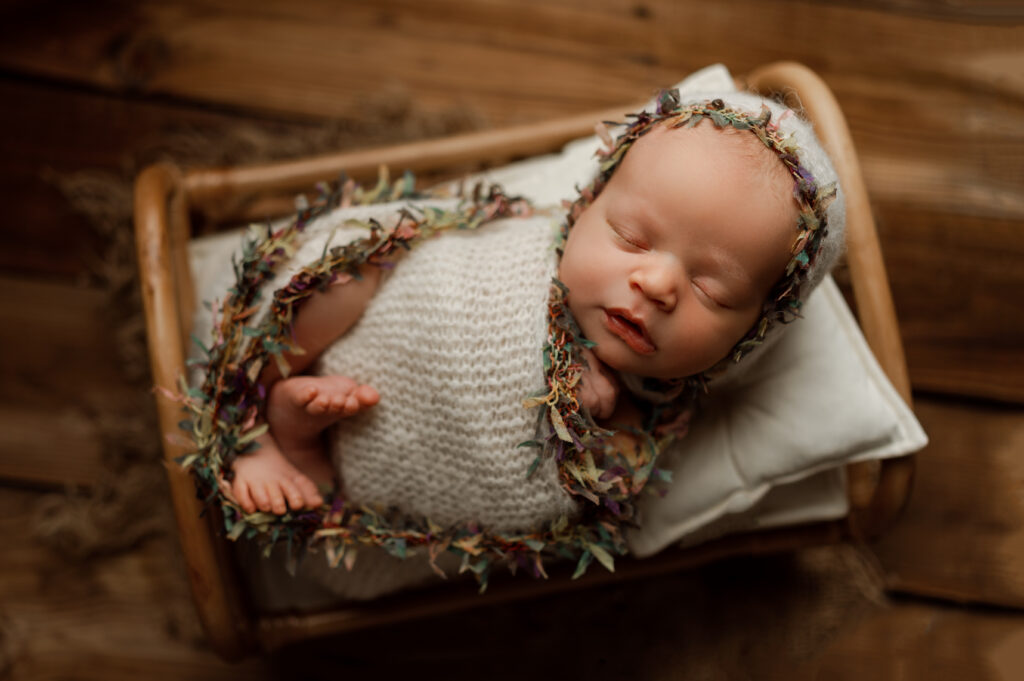 Newborn baby girl wrapped in a knitted wrap with colorful fringe and wearing a bonnet. She is posed on a bed that is sitting on a wooden floor. 