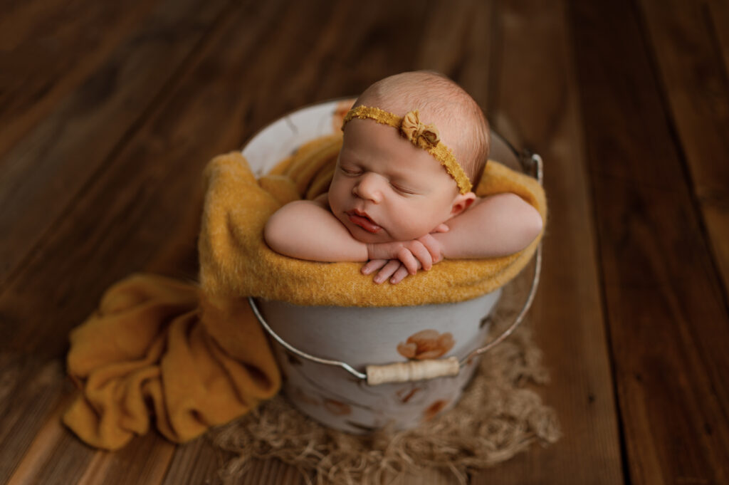 Newborn baby girl posed in a floral bucket. Color theme is yellow and she is on a wooden floor drop. 