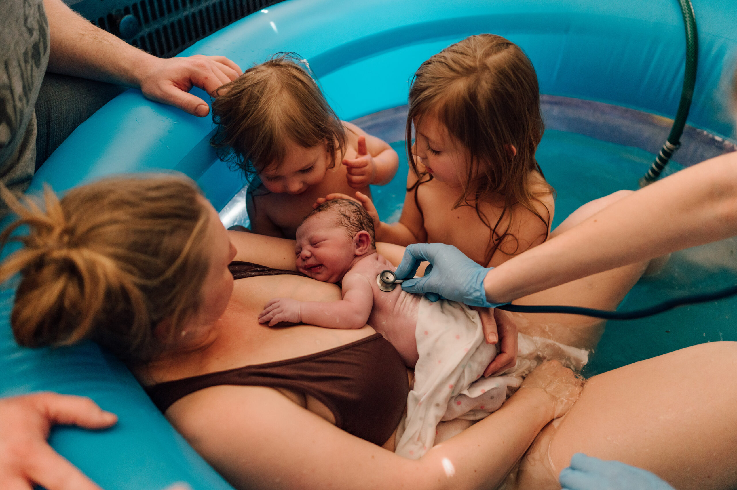 Birthing mother in pool shortly after delivery surrounded by her two other daughters and birth team.