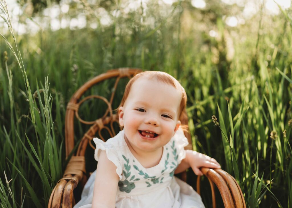 A beautiful red haired baby girl sitting in chair surrounded by tall grass with golden lighting scattering from behind. She has a huge beautiful grin and you can see her two top teeth.