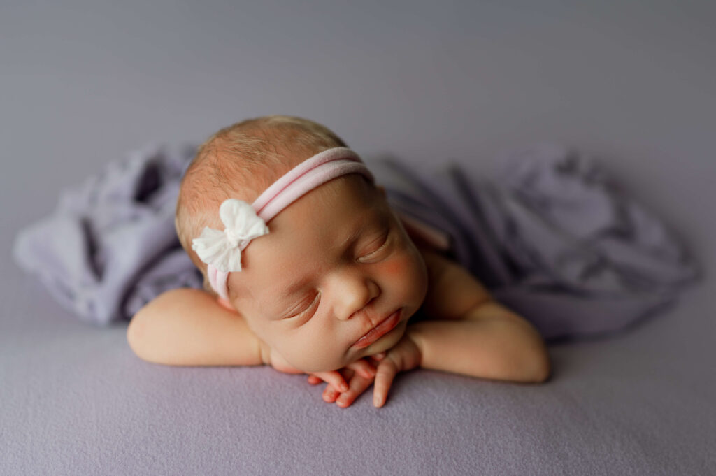 newborn girl posed facing forward on a purple backdrop carefully posed specifically for newborn photography with a headband on