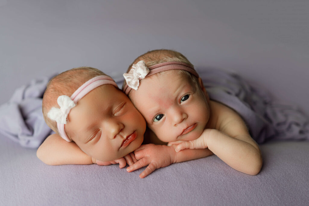 twin newborn girls posed facing forward on their belly snuggling close with one twin sleeping and the other awake looking at the camera on a purple backdrop.