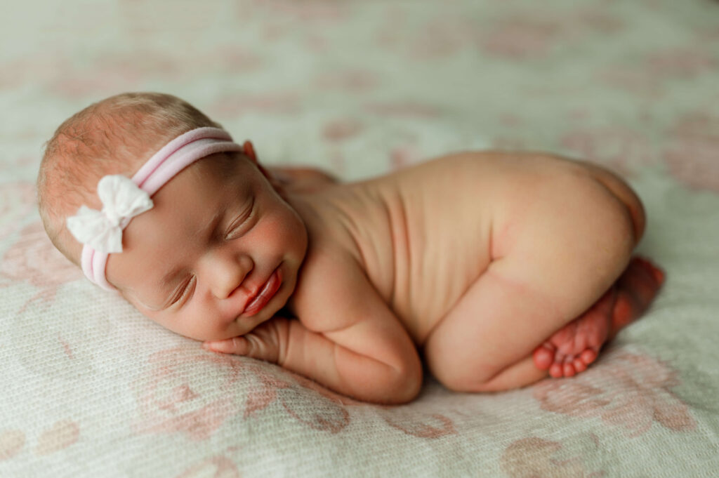 newborn girl posed on a floral soft pink and white backdrop in a newborn photography specific pose. baby is naked.