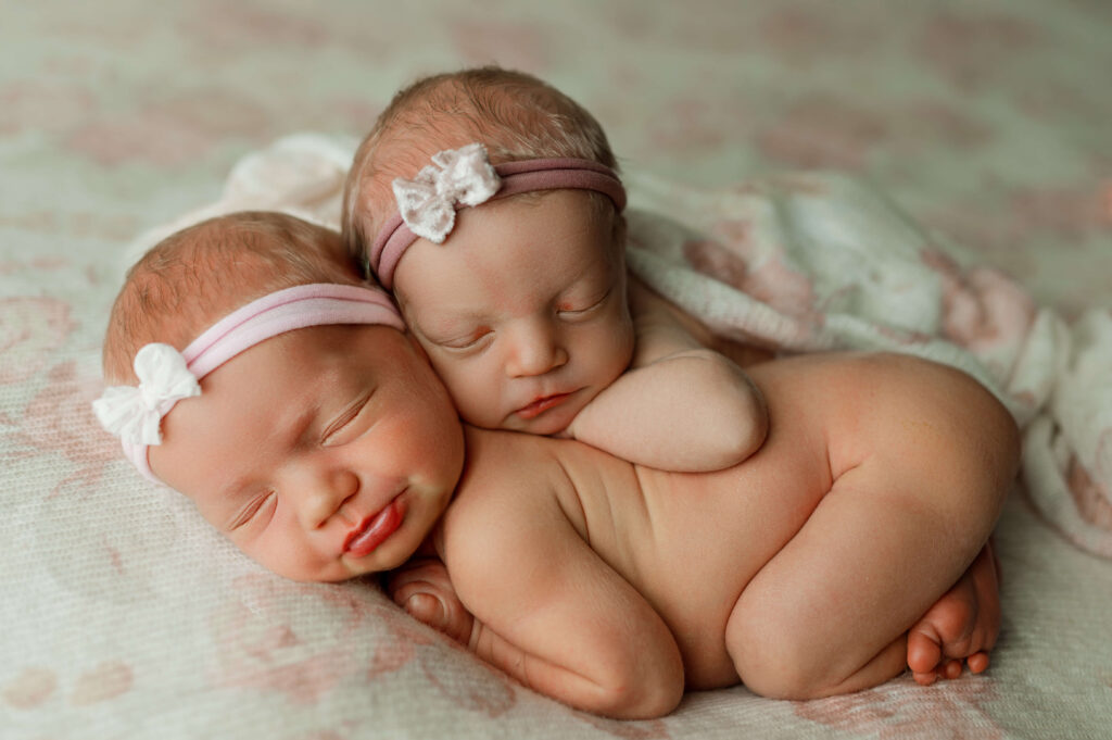 twins newborn girls posed on soft white and pink floral backdrop with one girl posed tastefully and artistically on top of the other twin.