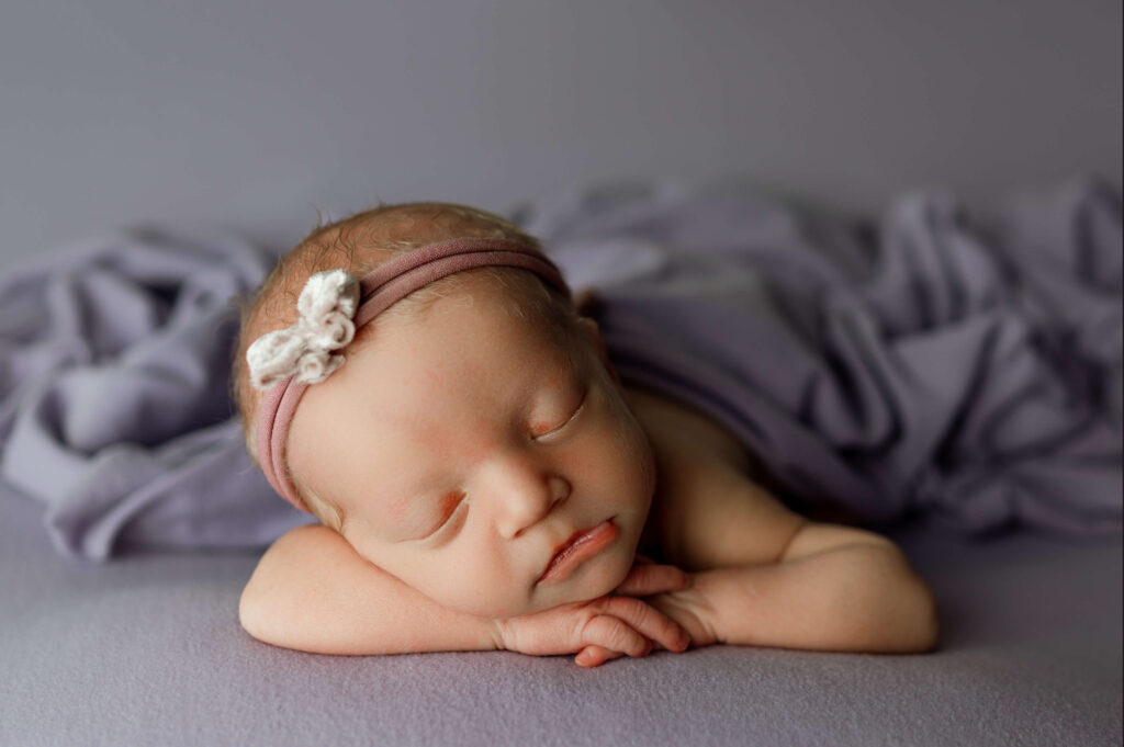 newborn girl posed facing forward on a purple backdrop carefully posed specifically for newborn photography with a headband on too
