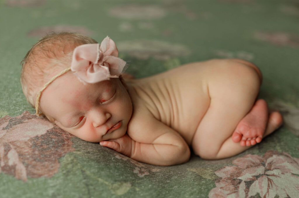 newborn girl posed on a floral green and pink backdrop in a newborn photography specific pose. baby is naked and has a large pink bow