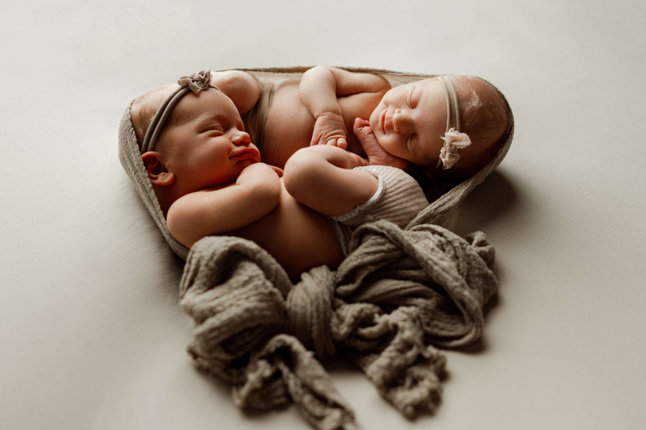 twins girls posed together facing eachother laying on a tan backdrop mimicing the womb. they are cuddled together and look content. This is a newborn photographer photograph so it is artful and safely done.