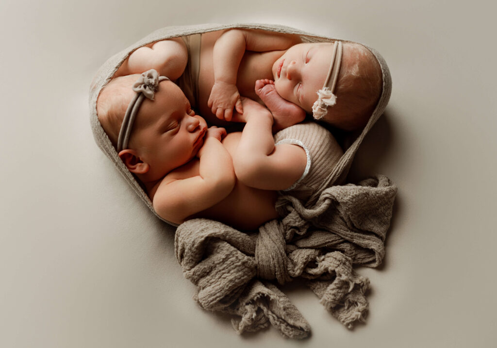 twins girls posed together facing eachother laying on a tan backdrop mimicing the womb. they are cuddled together and look content. This is a newborn photographer photograph so it is artful and safely done.