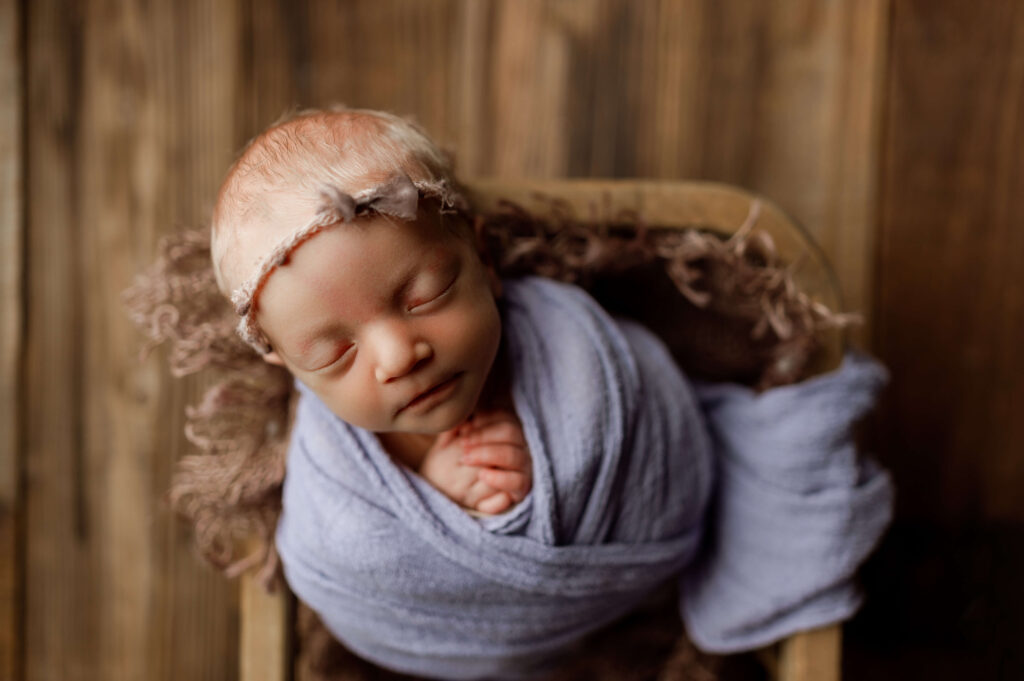 baby girl wrapped in purple place in a basket posed specifically for newborn photography with a headband on