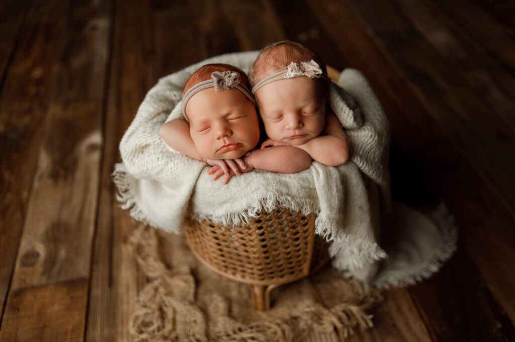 twin baby girls posed in a whicker basket together facing forward on the edge of the basket.