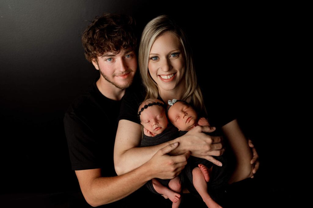 mom holding newborn twin girls in black wraps against a black backdrop with dad embracing mom and babies from the side. everyone is looking at the camera smiling
