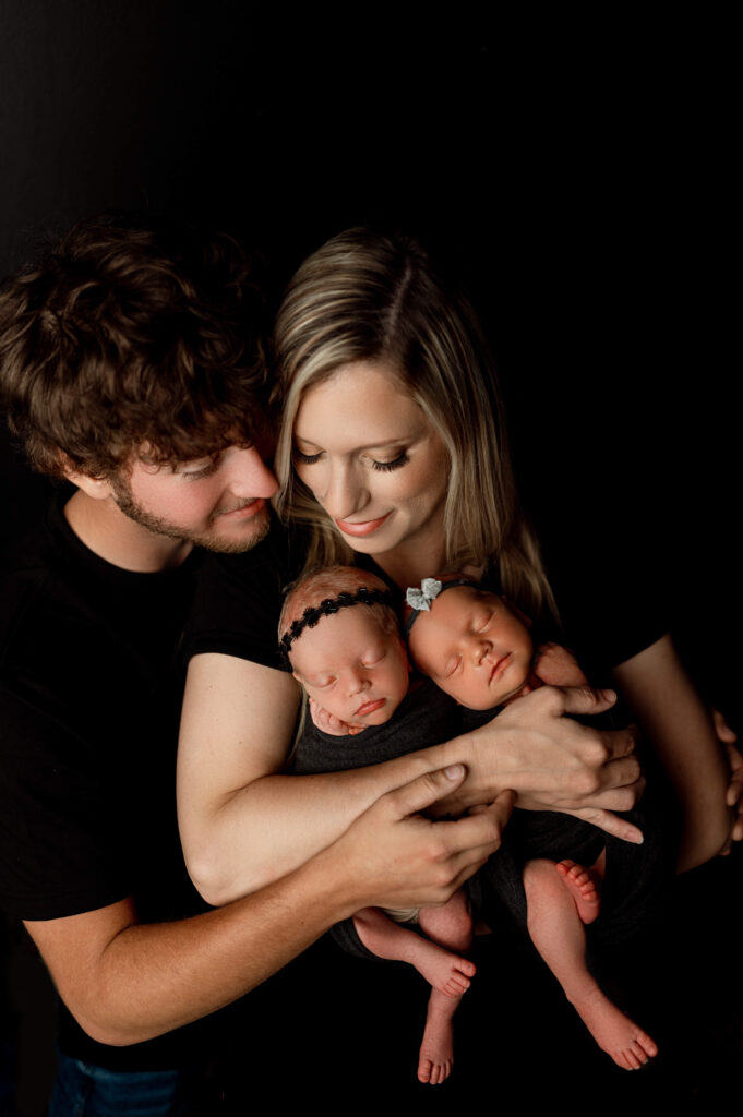 mom holding newborn twin girls in black wraps against a black backdrop with dad embracing mom from the side snuggling in to mom and babies