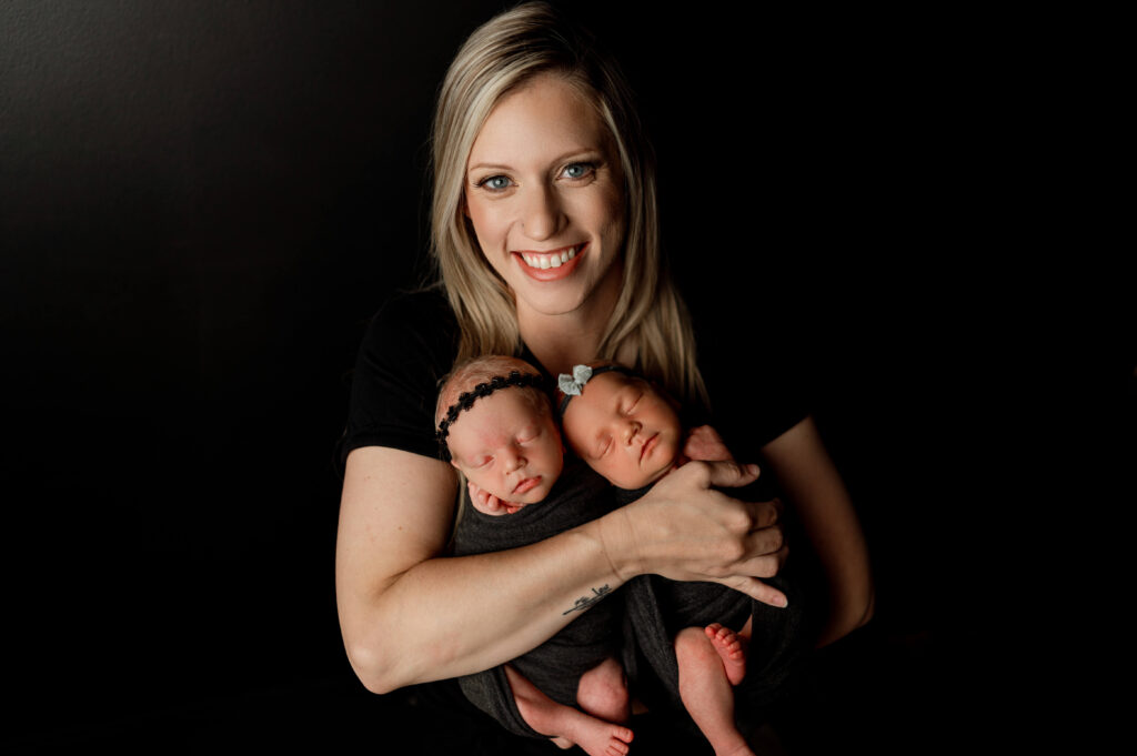 mom holding newborn twin girls in black wraps against a black backdrop