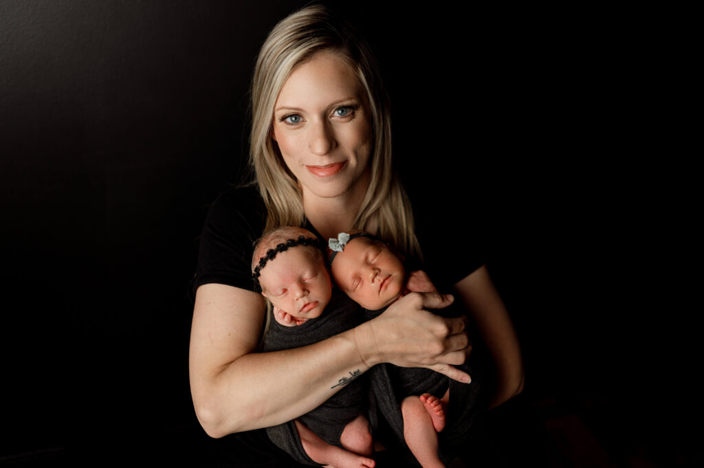 mom holding newborn twin girls in black wraps against a black backdrop in newborn studio setting