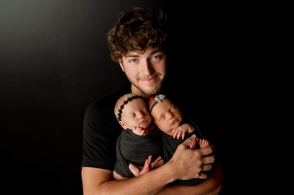 dad holding newborn twin girls in black wraps against a black backdrop