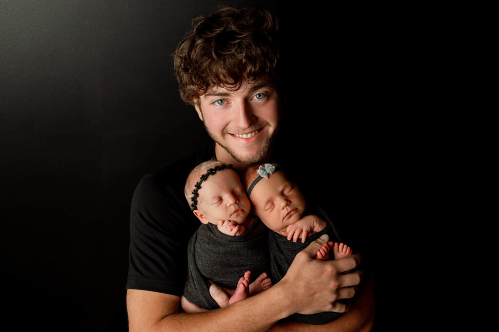 dad holding newborn twin girls in black wraps against a black backdrop in newborn studio with professional lighting