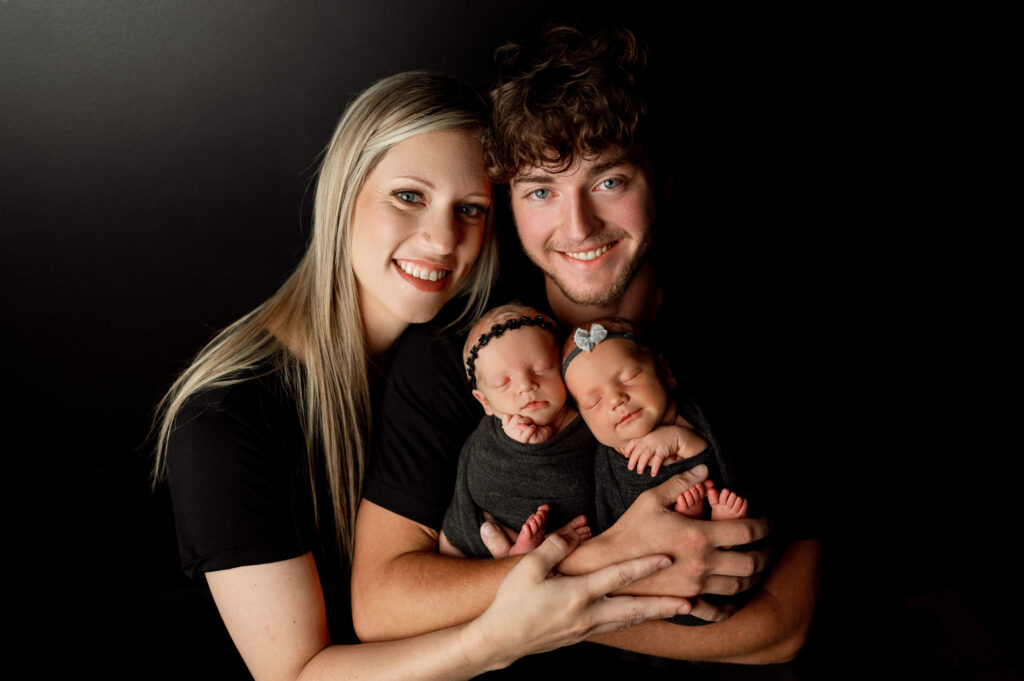dad holding newborn twin girls in black wraps against a black backdrop with mom embracing dad and babies from the side. everyone is looking at the camera smiling