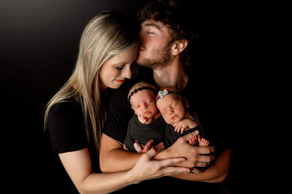 dad holding newborn twin girls in black wraps against a black backdrop with mom embracing dad and babies from the side. everyone is snuggled close and dad is kissing mom