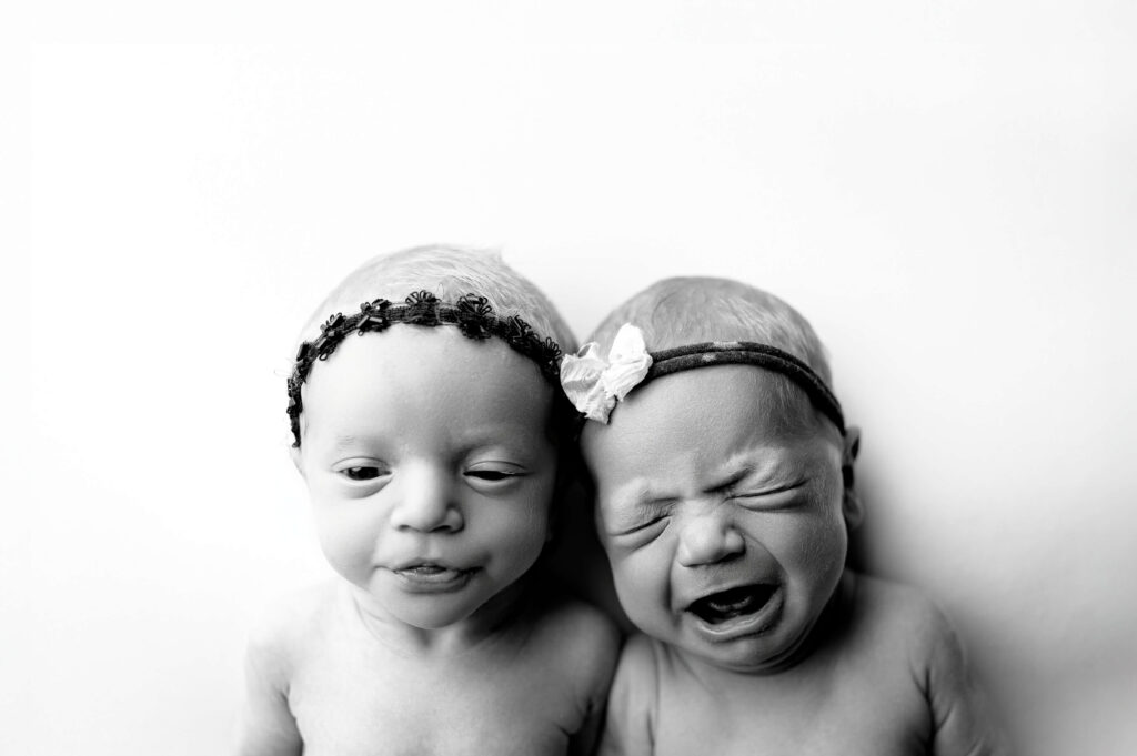 twin newborns laying on their back on a white backdrop looking up to camera making cute expressions to the camera