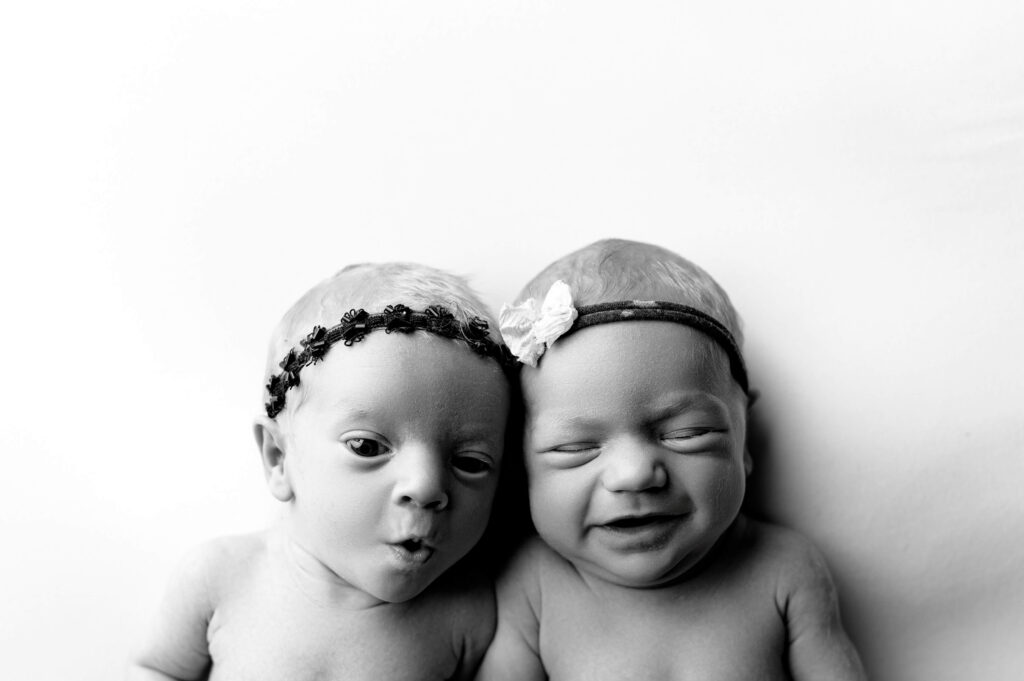 twin newborns laying on their back on a white backdrop looking up to camera making cute expressions to the camera