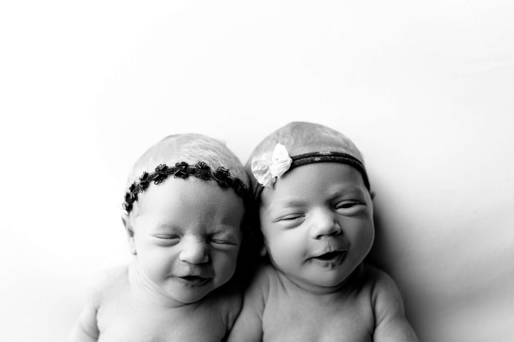 twin newborns laying on their back on a white backdrop looking up to camera making cute expressions to the camera