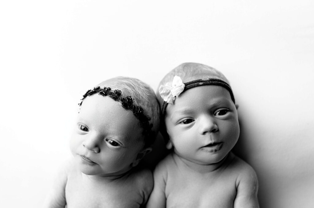 twin newborns laying on their back on a white backdrop looking up to camera making cute expressions to the camera