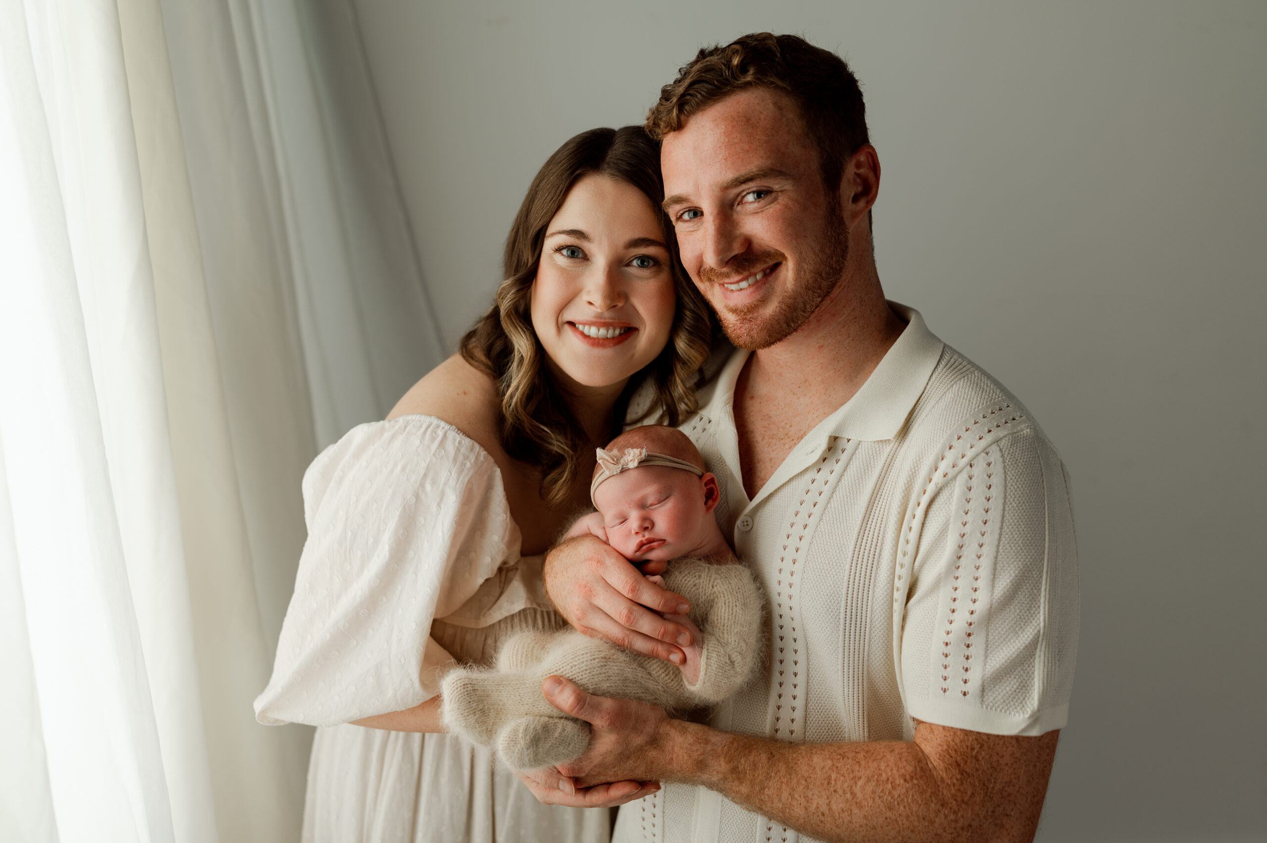 An in studio newborn portraits of a mom and dad with their baby. Dad is holding baby and mom is hugging them and their smiling at the camera