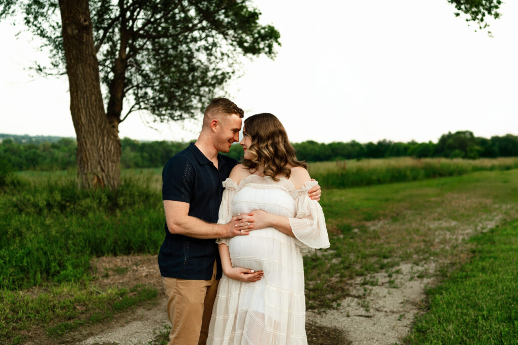 a mom and dad posing outdoors for maternity photos in a white flowy dress