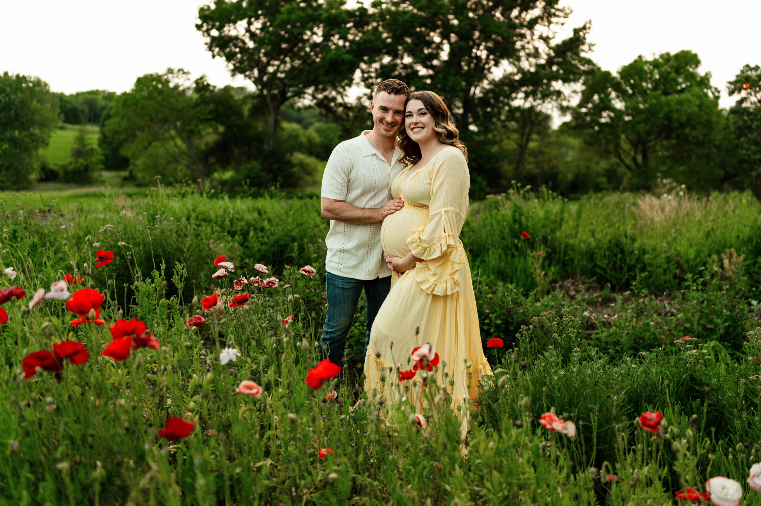a couple posing for maternity portraits surrounded by red flowers. mom is in a yellow dress and dad is in a cream shirt